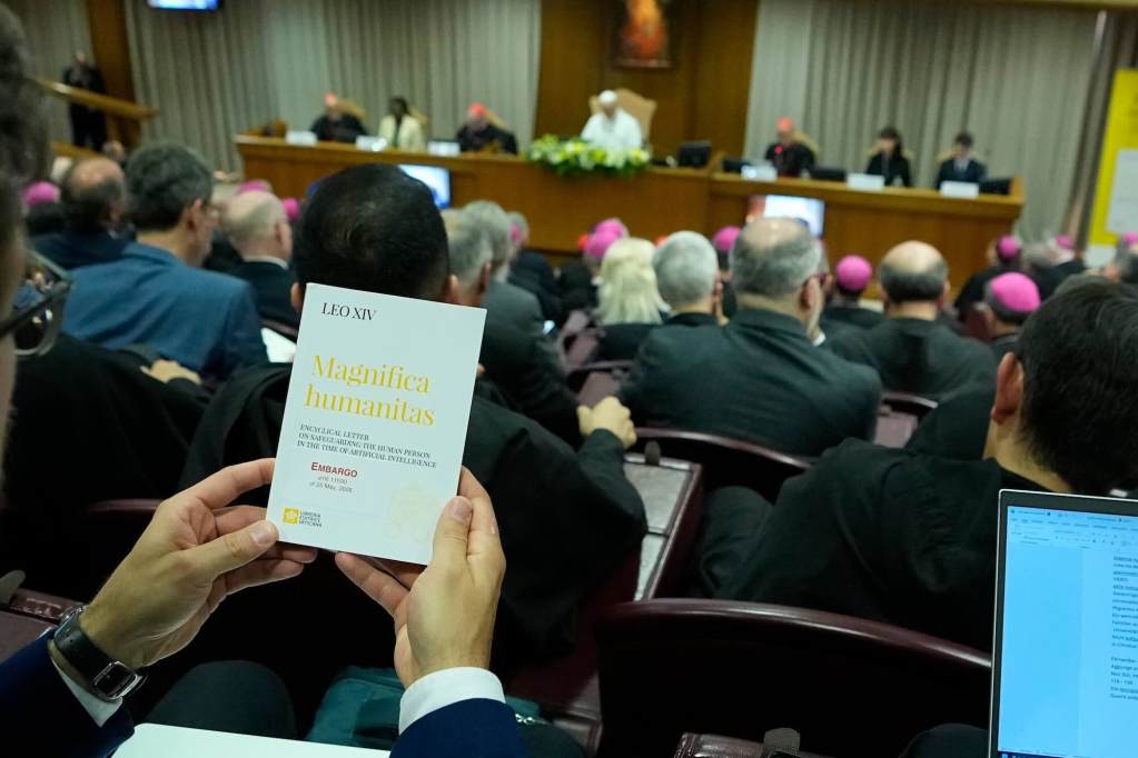 A man holds a card that says “Magnifica humanitas” while seated in an auditorium. Pope Leo XIV is seen seated on the stage.