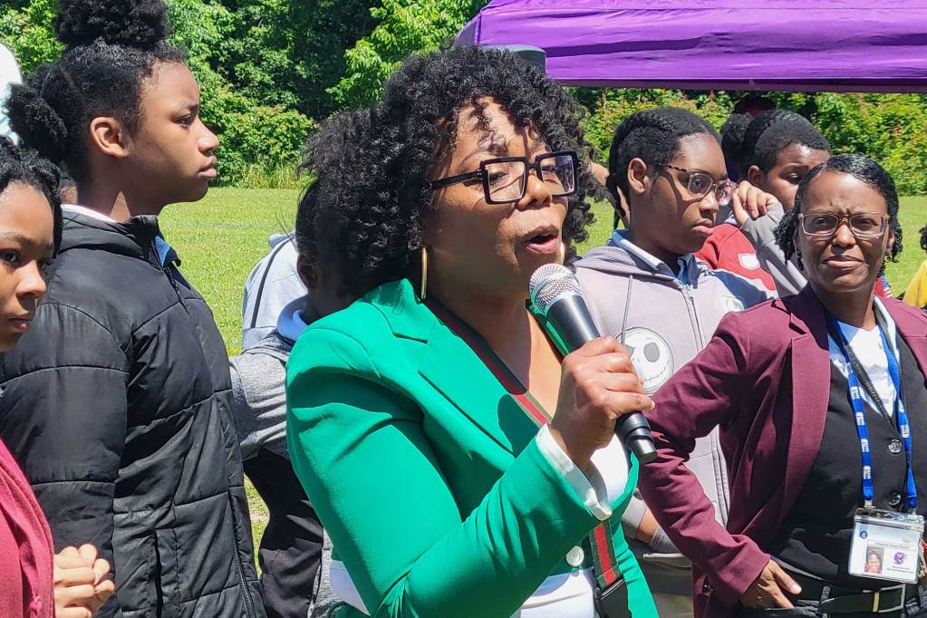 A woman in a green jacket speaks into a mic at an outside event