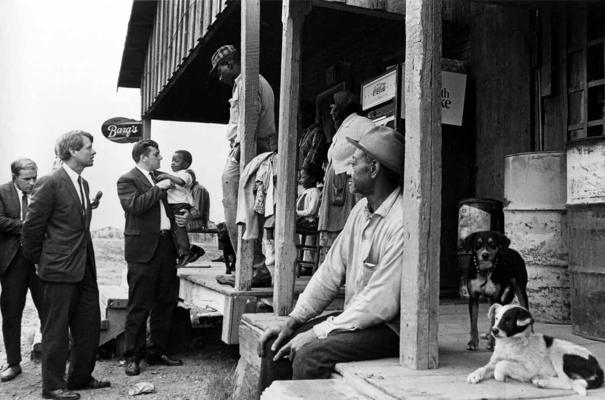 An old black and white photo from the 1960's showing Robert F. Kennedy and other white men in suits visiting outside a country store