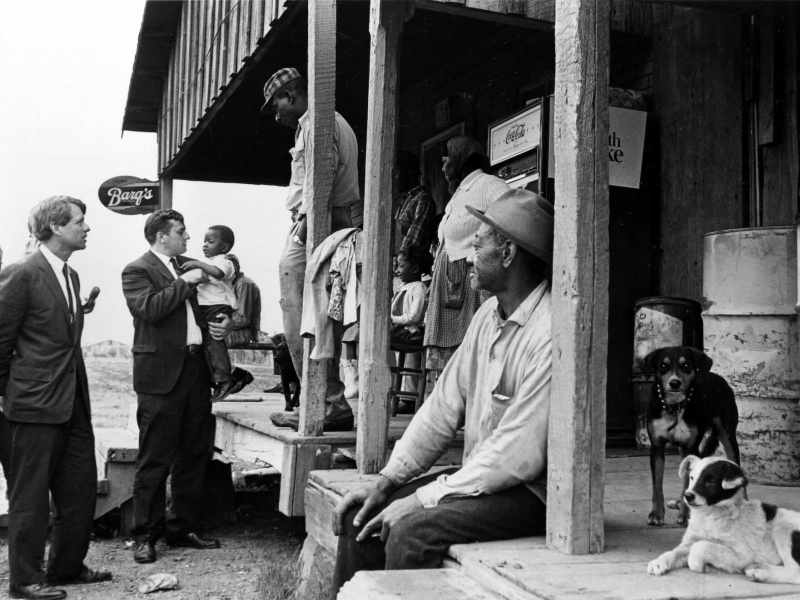 An old black and white photo from the 1960's showing Robert F. Kennedy and other white men in suits visiting outside a country store