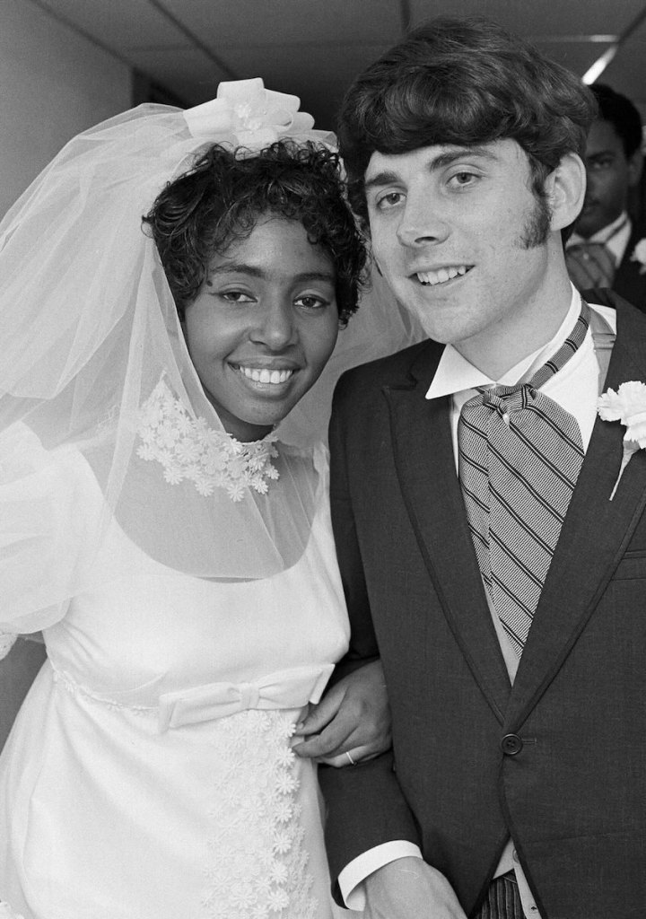 An old black and white photo from the 1970's of a Black woman in a white wedding dress and a White man dressed as a groom, smiling together