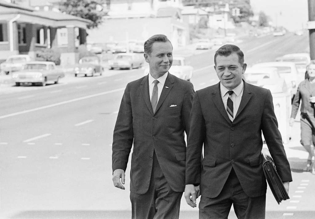 An old black and white photo from the 1960's of two white men in suits walking down a sidewalk