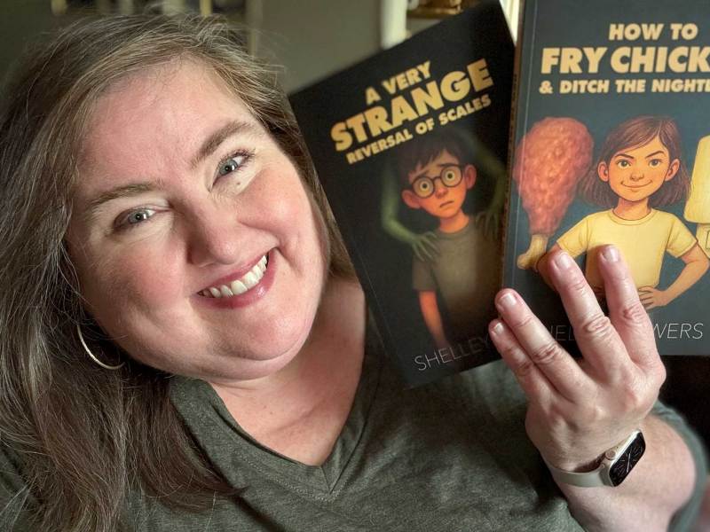 A woman with long brown hair smiles while holding up two books with illustrations on the covers. The titles are "A Very Strange Reversal of Scales" and "How to Fry Chicken & Ditch the Nightlight"
