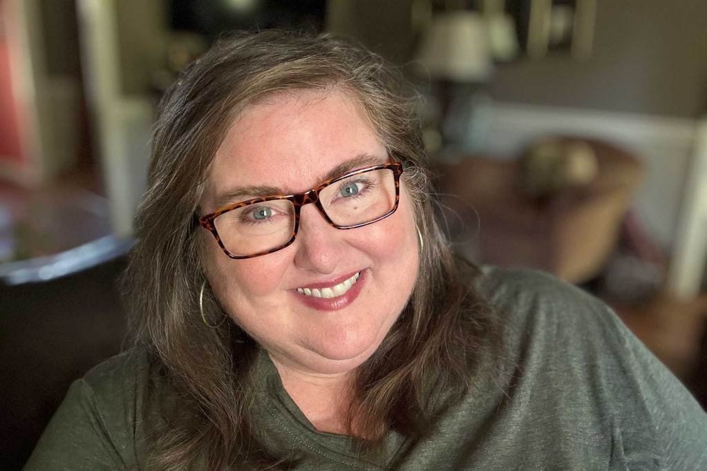 A woman with long brown hair and tortoise shell glasses smiles inside her home