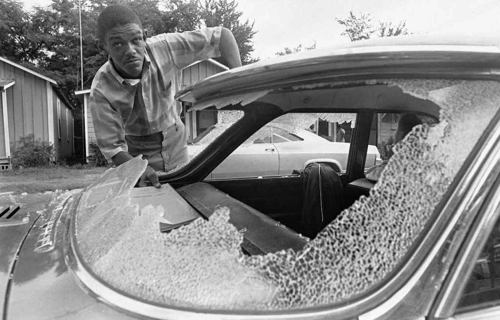 An old black and white photo from the 1960's of a man inspecting the damage of a smashed out rear car window