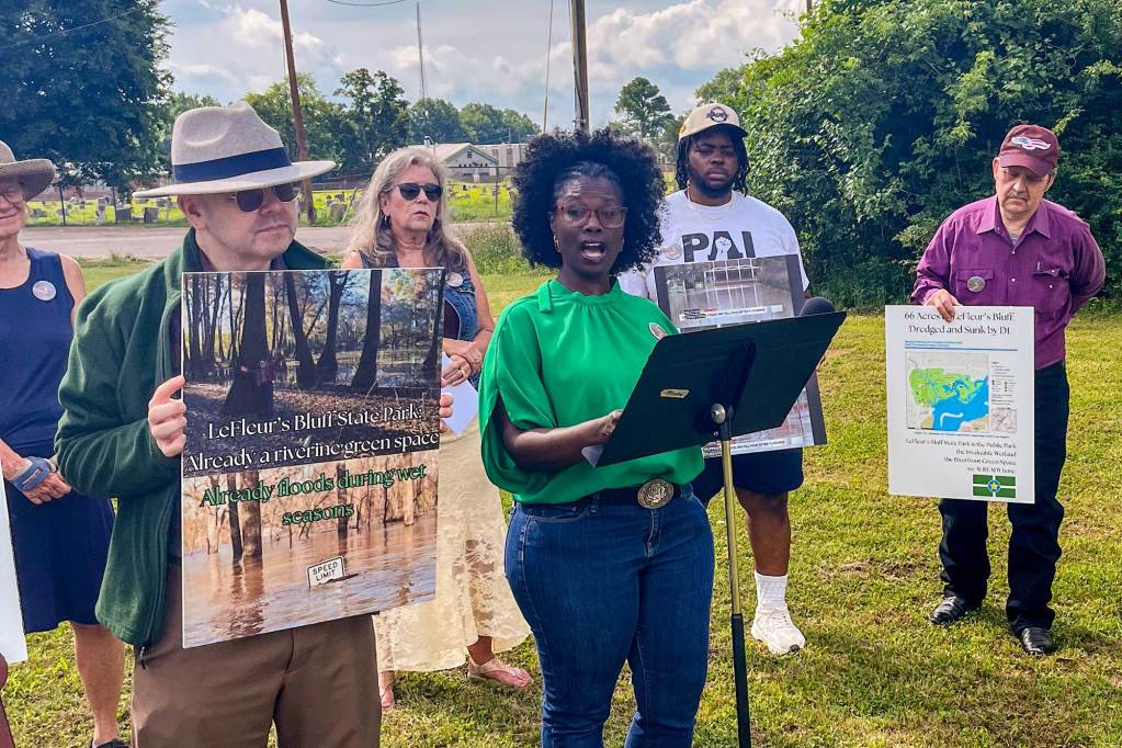 A woman in a green top speaks at an outdoors press conference, as others behind her hold up signs