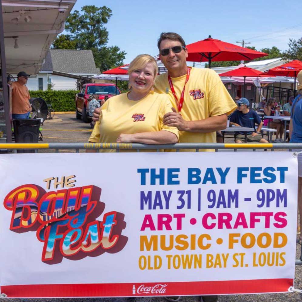 Two people in matching Bay Fest shirts stand behind a banner for The Bay Fest being held on May 31