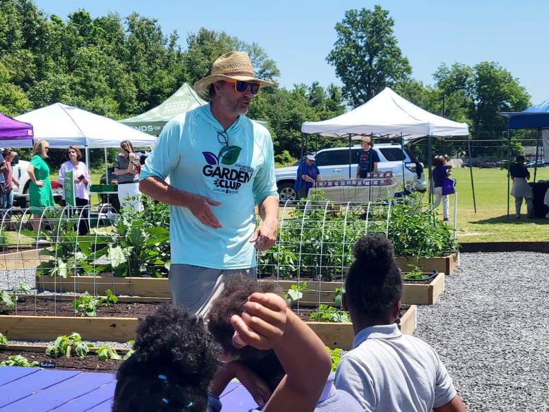 A man in a Garden Club tshirt speaks to students about the raised gardens beside them