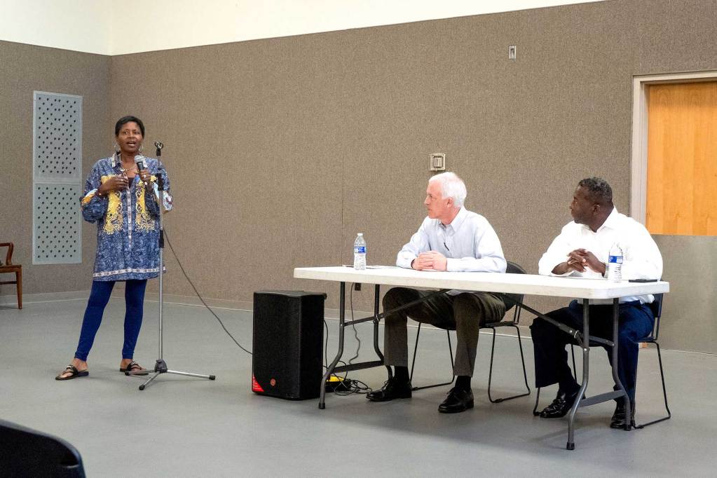 David Blount and Chris Bell sit at a table watching a woman speak into a microphone.