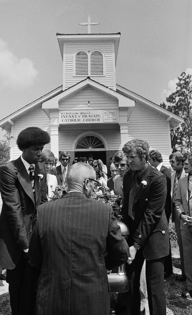 An old black and white photo from the 1970's showing pallbearers moving a casket in front of a white church