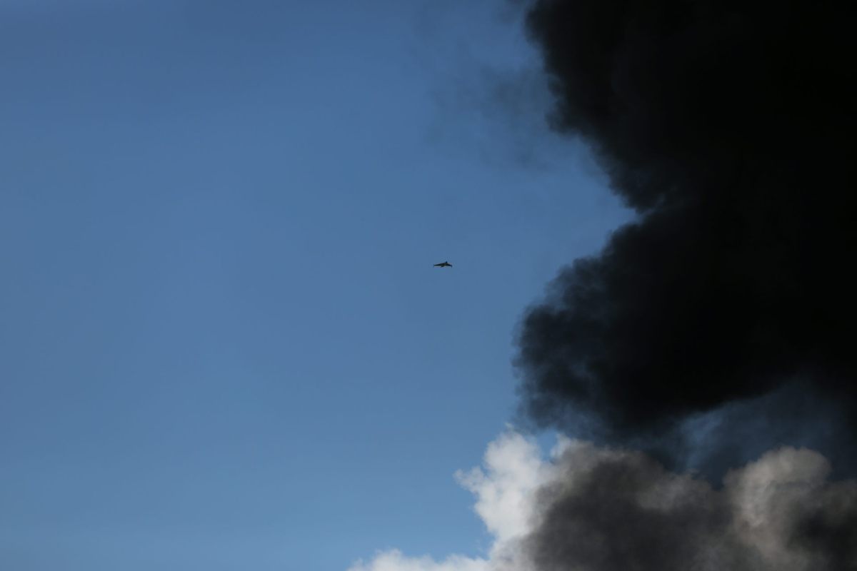 A drone flying high in the sky over a black plume of smoke.