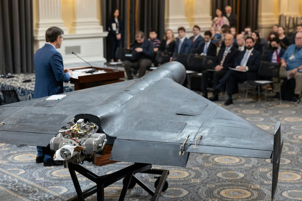 A drone vehicle on display in a conference room with a view behind Mike Johnson, who is speaking at a podium in front of a crowd of people.