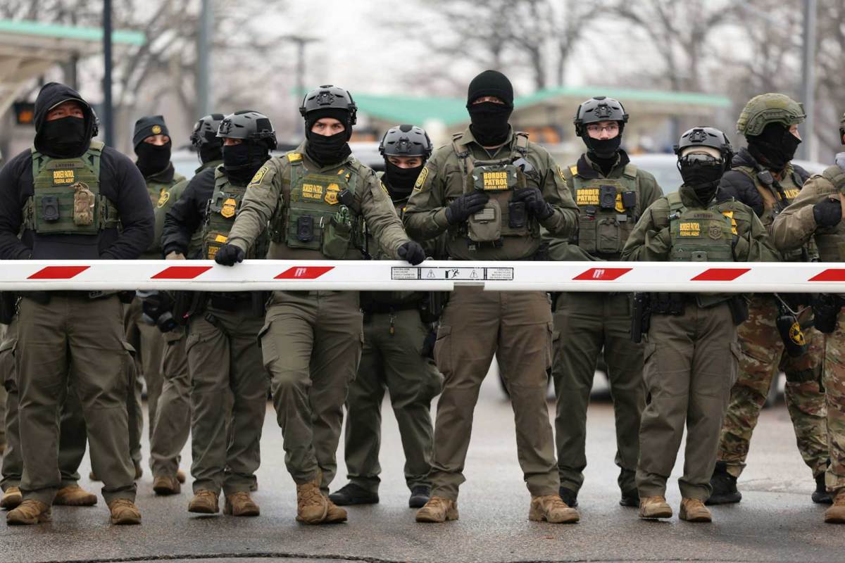 Several border patrol agents wearing vests and masks behind a crossing barrier.