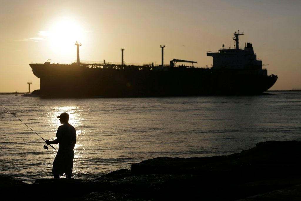 A man fishing on the shore with a large oil tanker vessel on the sea in the background against the sunset.