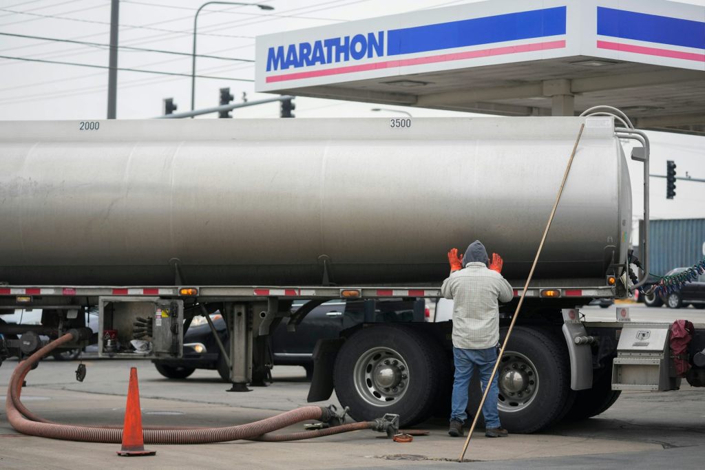 A man stands near a gas tanker truck. A long nozzle connects the tank to an outlet on the ground.