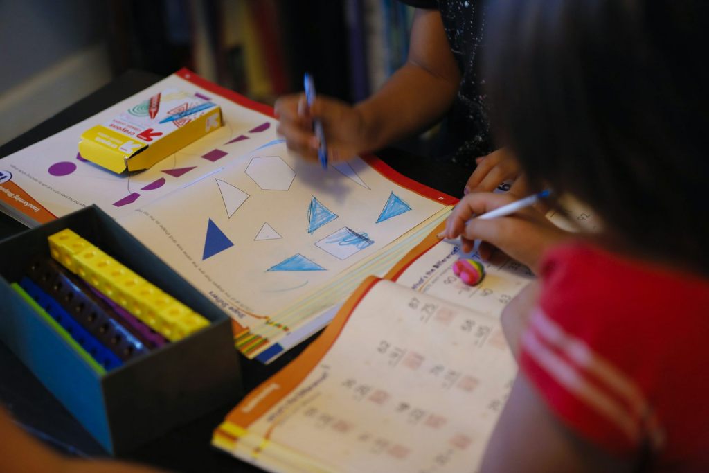 Two children, whose arms and shoulders are seen in this cropped photo, hold pens and lean over workbooks, one of which has photos of triangles on it.