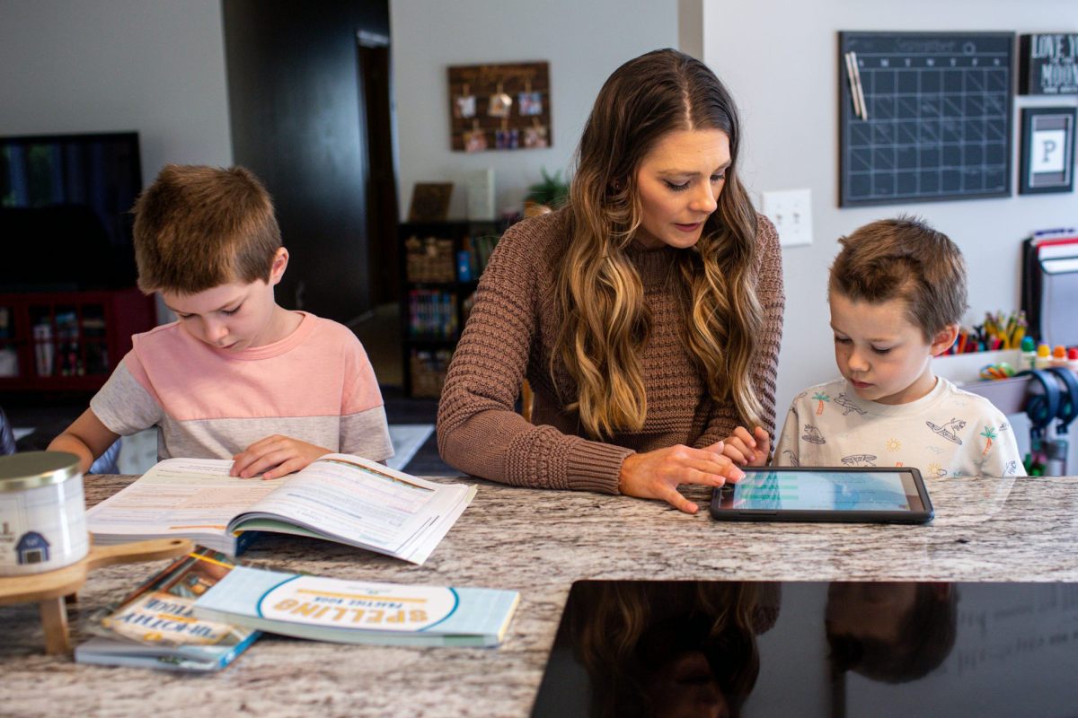 A woman seated between two young boys at a table, reading books and tablets
