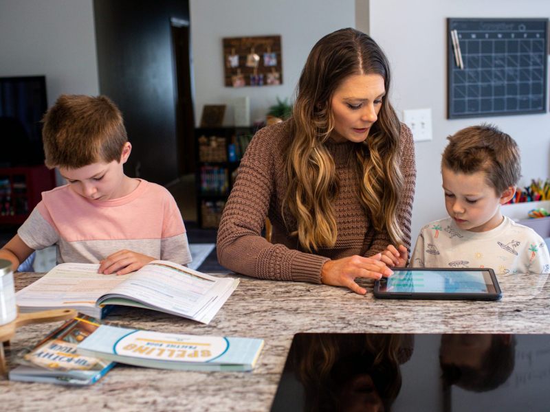 A woman seated between two young boys at a table, reading books and tablets