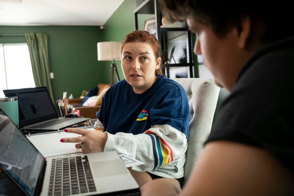 A woman wearing a long-sleeve shirt holds two fingers up near a laptop, as a teenage boy looks at the laptop and sits next to her.