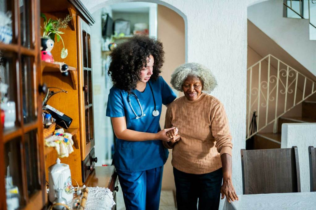 A woman wearing medical scrubs helping an older woman walk through a home
