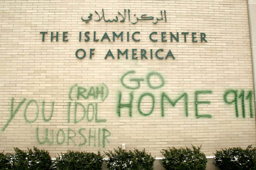 Lettering on a wall with Arabic text and below it “The Islamic Center of America” in English. Below it in graffiti are anti-Muslim phrases like “You Idol Worship” and “Go Home 911.”