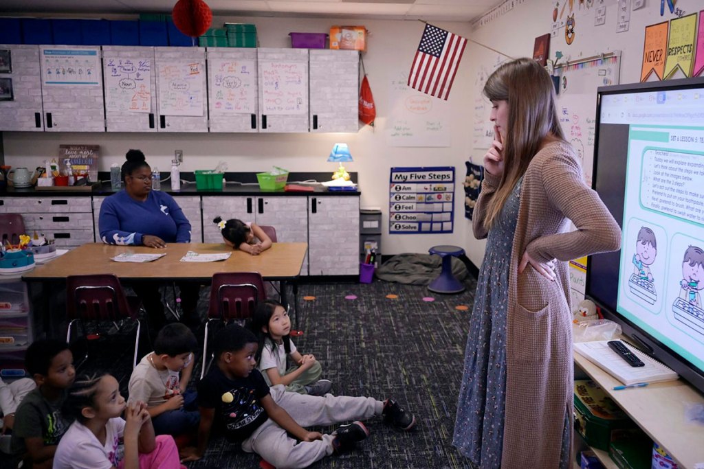 A woman stands near a projector screen in front of a group of children seated on the floor in a classroom.