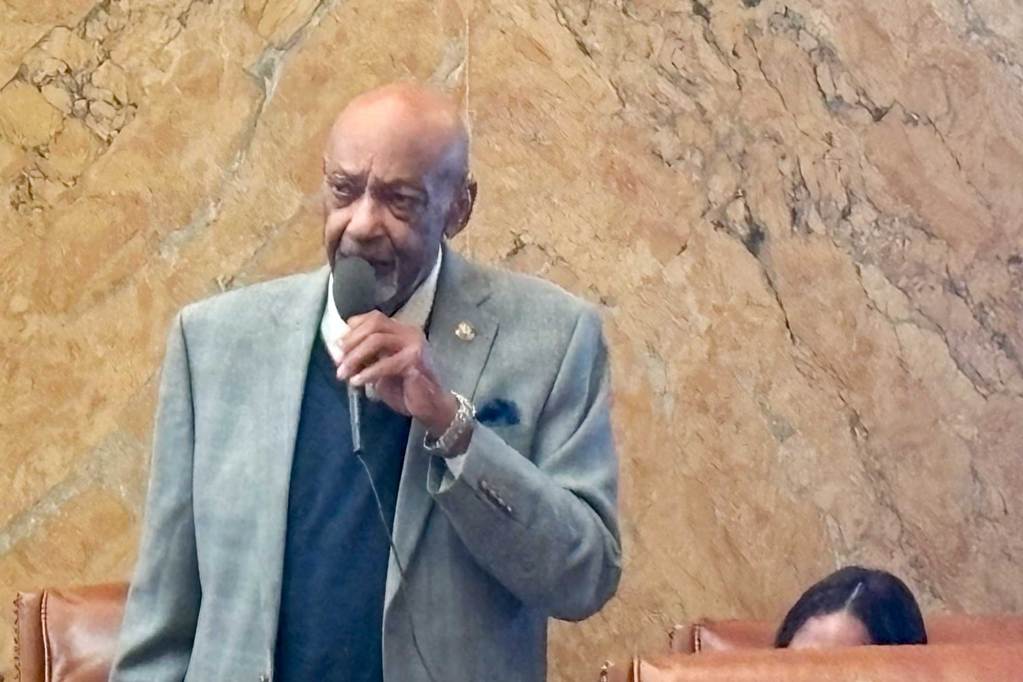 An older man in a suit stands up in a row of legislature desks and speaks into a mic