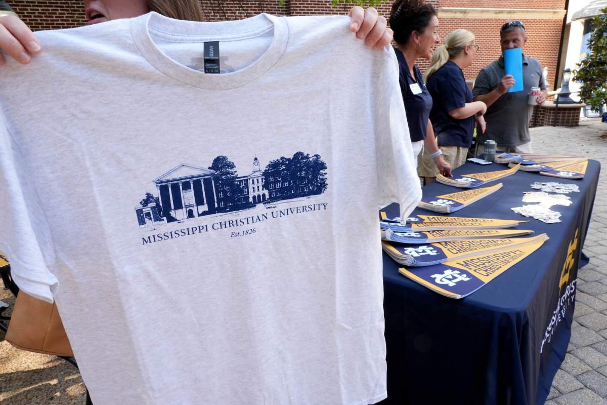 A person holds up a white tshirt that has a blue graphic of a college campus with the phrase “Mississippi Christian University.” Three other people are at a table behind them.