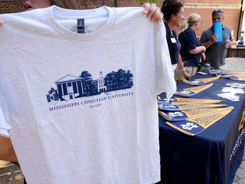 A person holds up a white tshirt that has a blue graphic of a college campus with the phrase “Mississippi Christian University.” Three other people are at a table behind them.