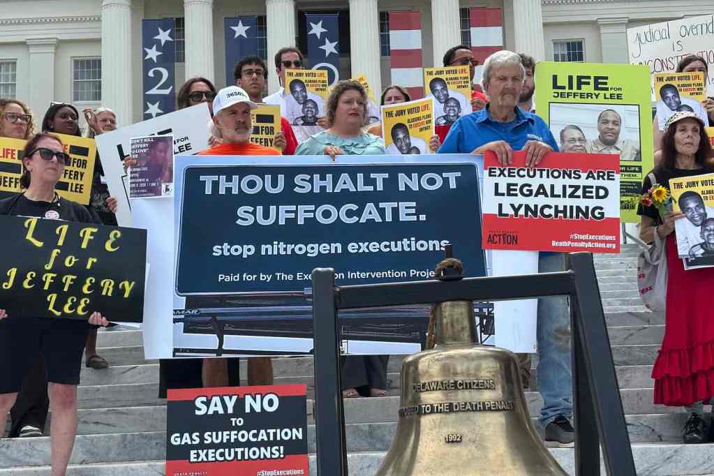 A sign that says “Thou Shall Not Suffocate. Stop Nitrogen Executions” displayed behind a bell and in front of several protestors holding signs.
