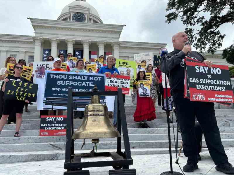 A man speaking into a mic next to a large bell. Several protestors holding signs are behind him on the steps of the Alabama Capitol Building.