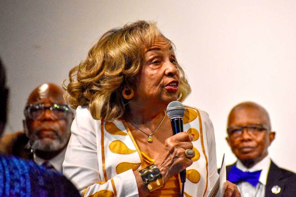 Flonzie Brown Wright, in a white and gold outfit, speaks with microphone in hand. Politicians sit behind her and listen