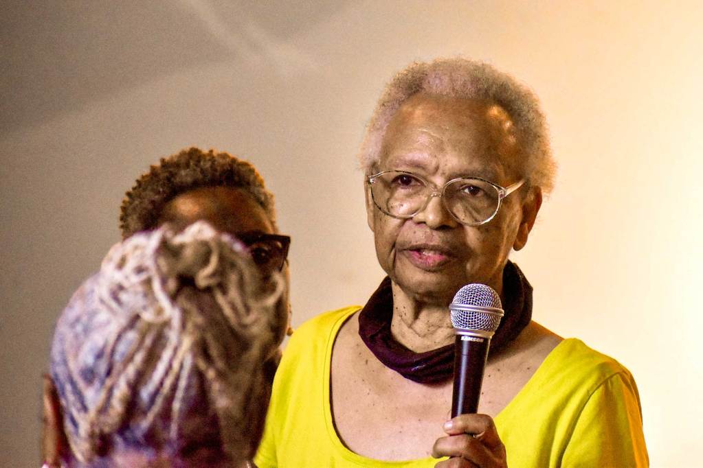 Hazel Hull, in a yellow outfit, holds a mic and speaks at an event as others listen