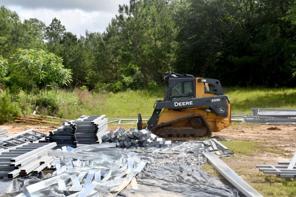 A yellow and black skid-steer loader sits near stacks of gray steel beams and metal strips at a grassy construction site.