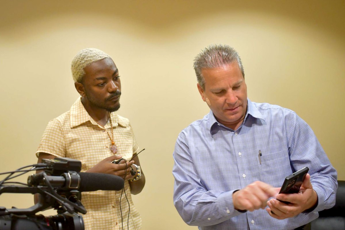 Two men stand in an office beside a video camera as one looks at a phone and the other holds cables.