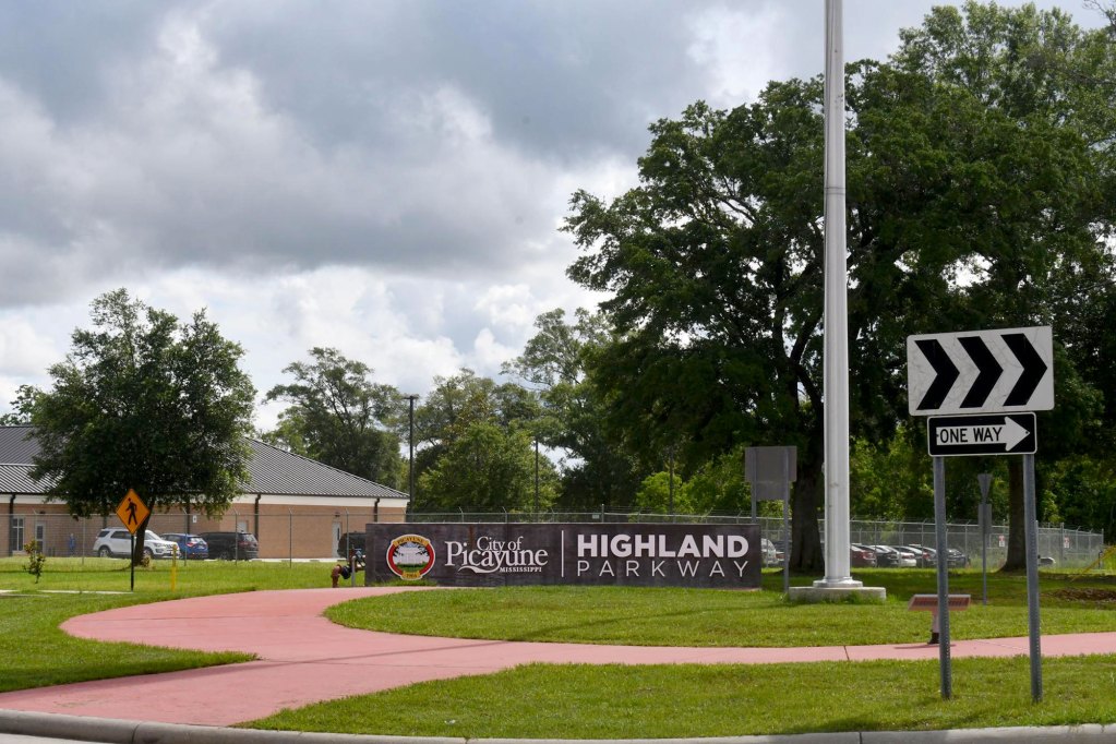 A Highland Parkway entrance sign, one-way arrow sign, sidewalk, grass and trees beneath a cloudy sky.