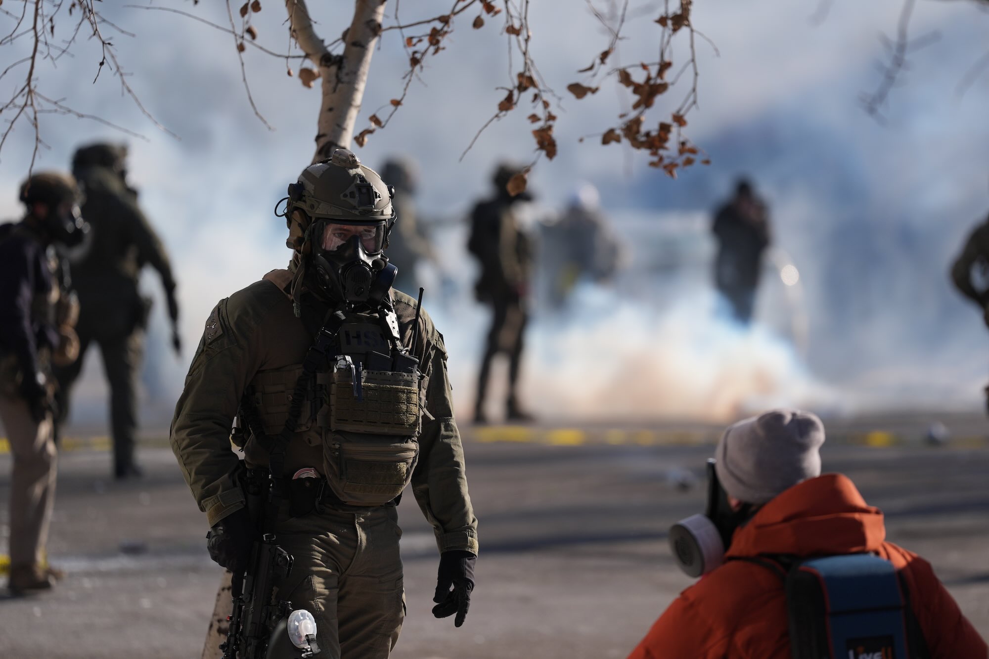 An immigration officer wearing protective gear standing in the street, with other officers and clouds of smoke in the background.