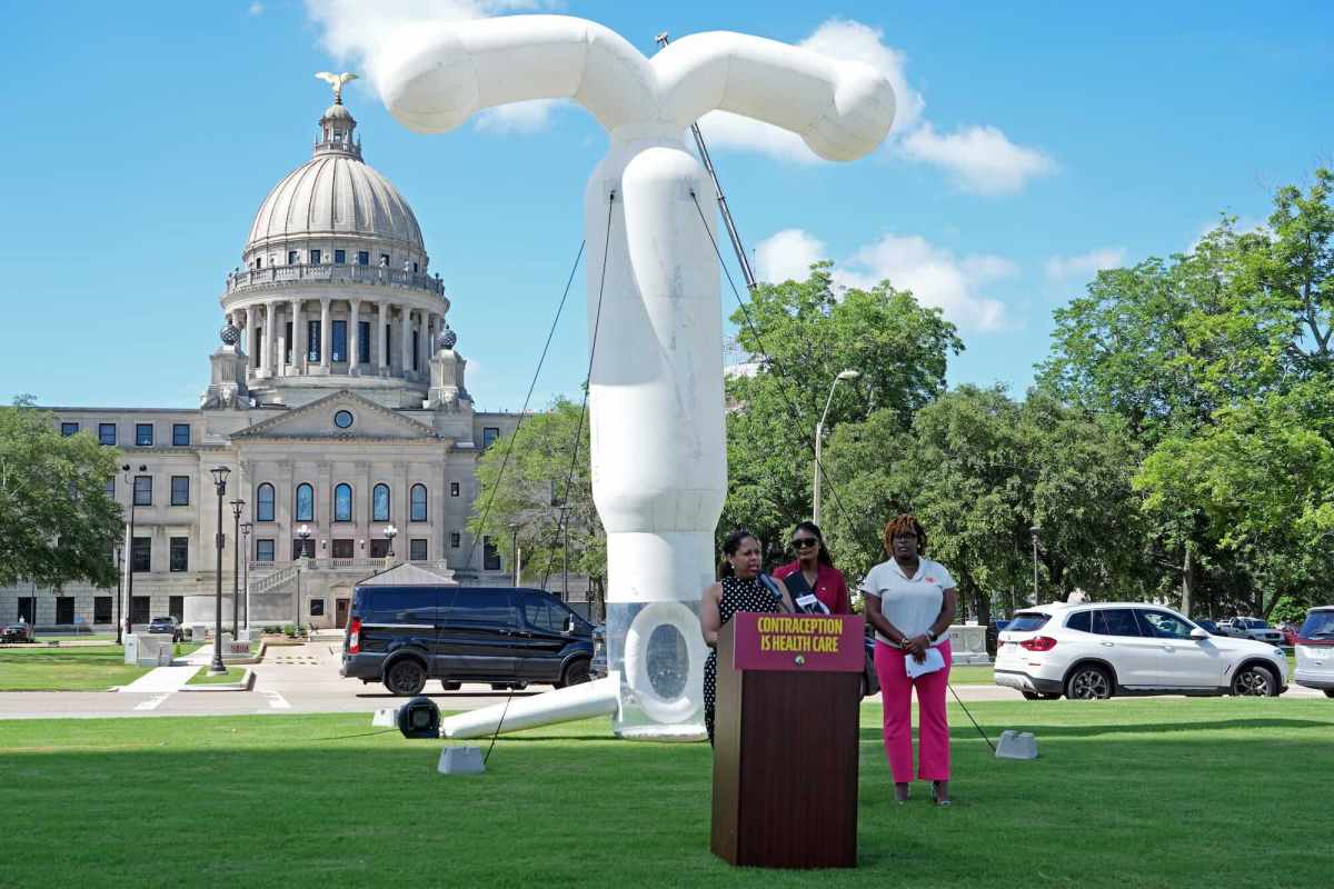 Three people speak at a podium on the green lawn in front of the Mississippi capital building. A large white inflated IUD stands behind the people.