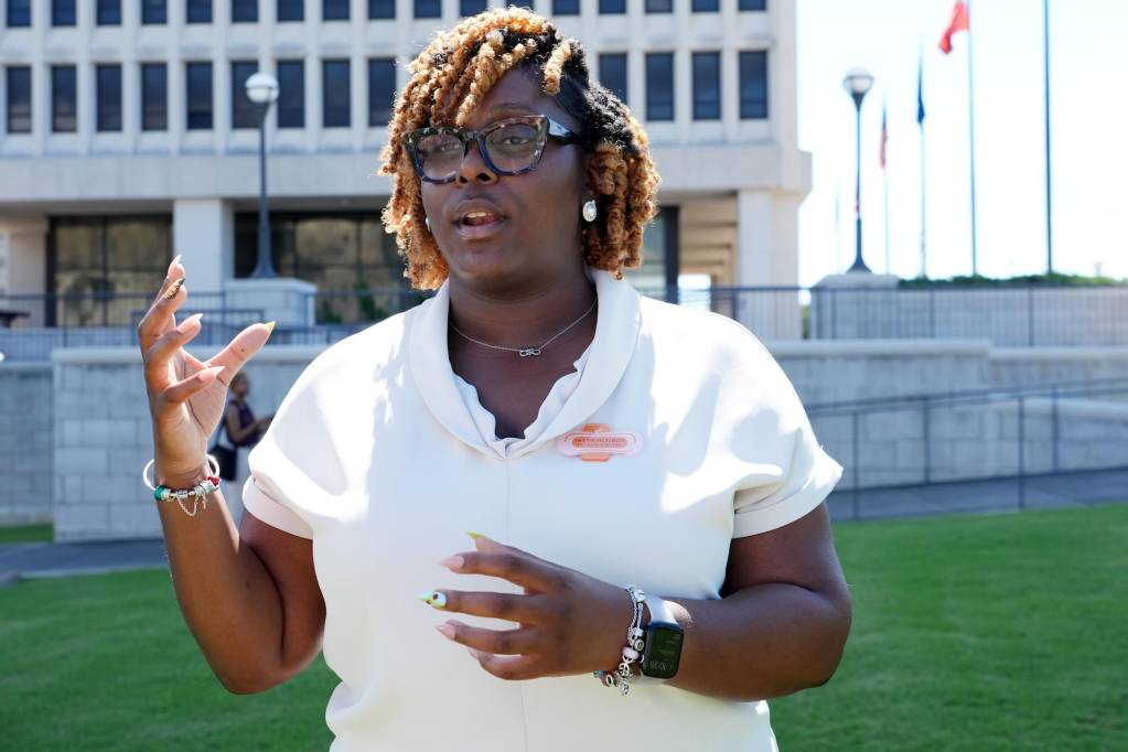 A woman with short hair and glasses speaks outside of a government building