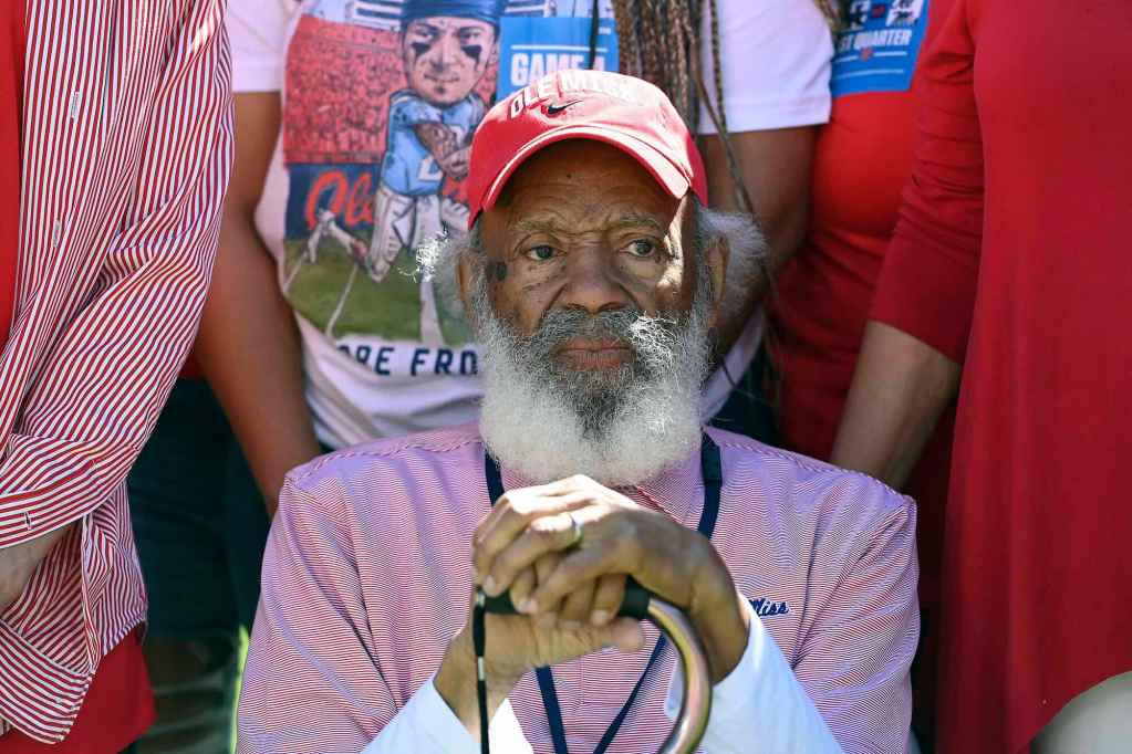 James Meredith, wearing a Ole Miss shirt and hat, sits leaning on his cane as others in Old Miss colors stand behind him