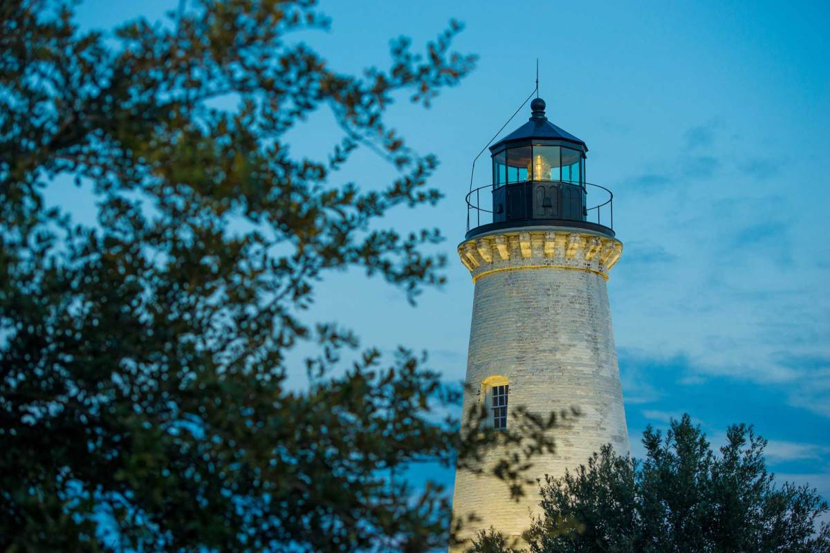 A white lighthouse seen past some greenery at dawn, still underlit by yellow lights.
