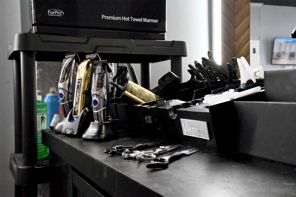 A black barber workstation holds several electric clippers, clipper guards, combs and shears, with a hot towel warmer above it.