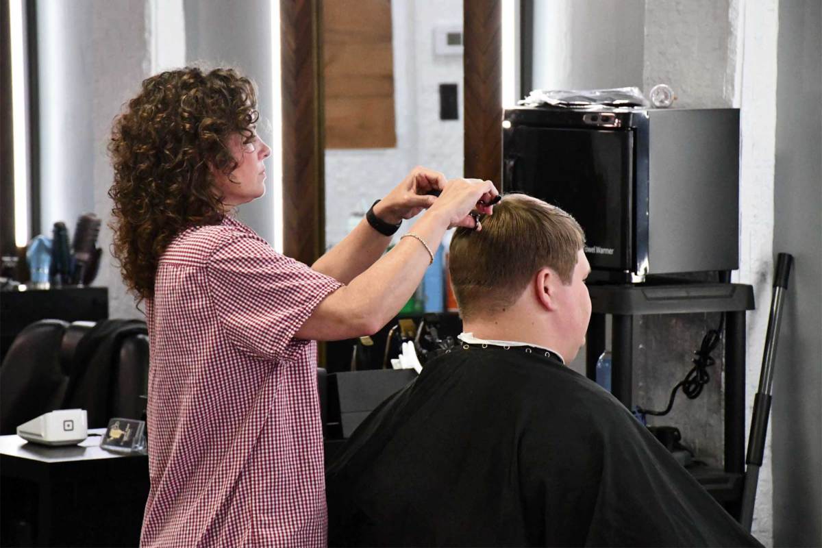 Crystal McCormick, wearing a red-and-white checkered shirt, trims the top of a seated client’s hair inside a barbershop.