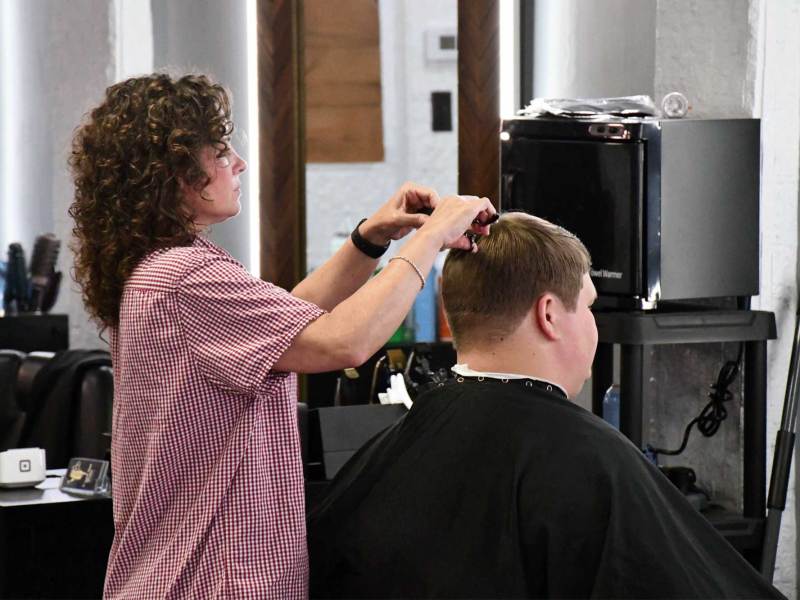 Crystal McCormick, wearing a red-and-white checkered shirt, trims the top of a seated client’s hair inside a barbershop.
