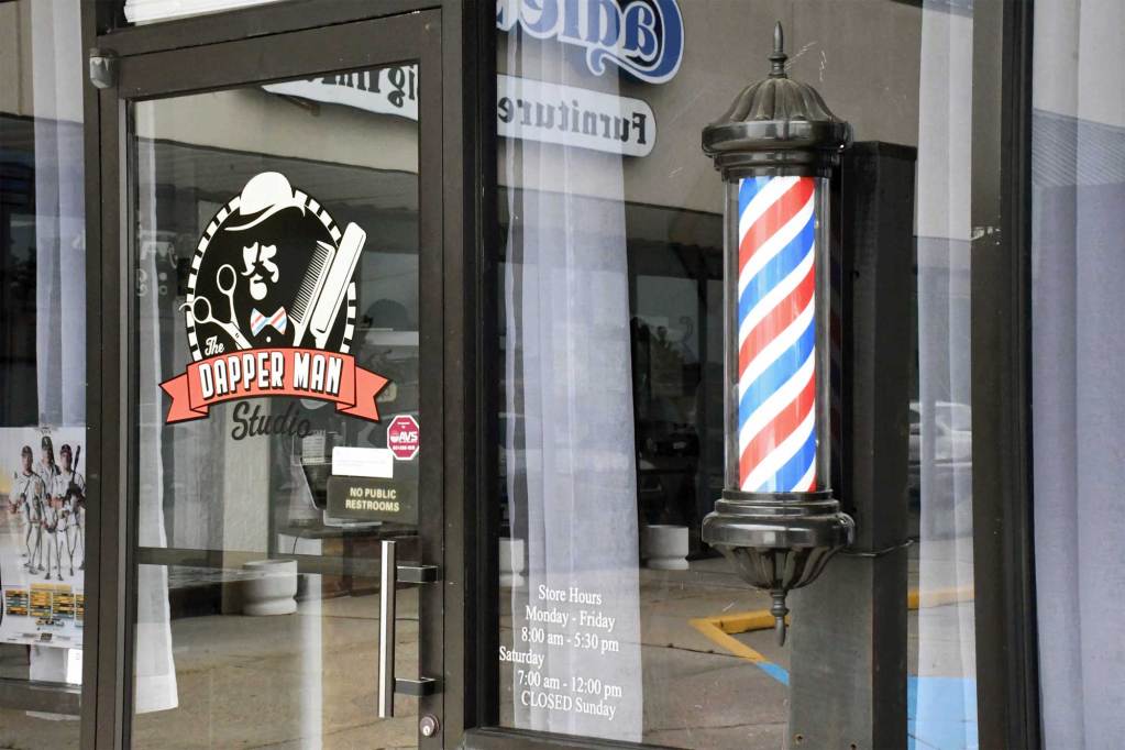 A glass storefront shows The Dapper Man Studio logo on the door and a red, white and blue barber pole mounted beside it.
