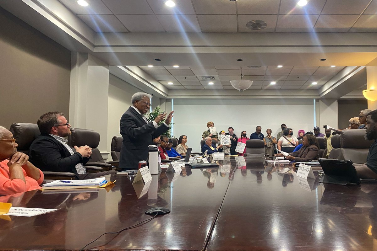 A meeting held with people sitting around a large conference table as others stand by the back wall