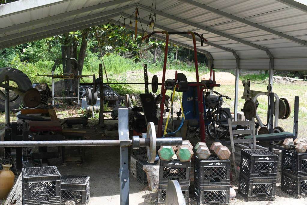 An outdoor home gym with weights, benches and exercise equipment set up under a metal carport.