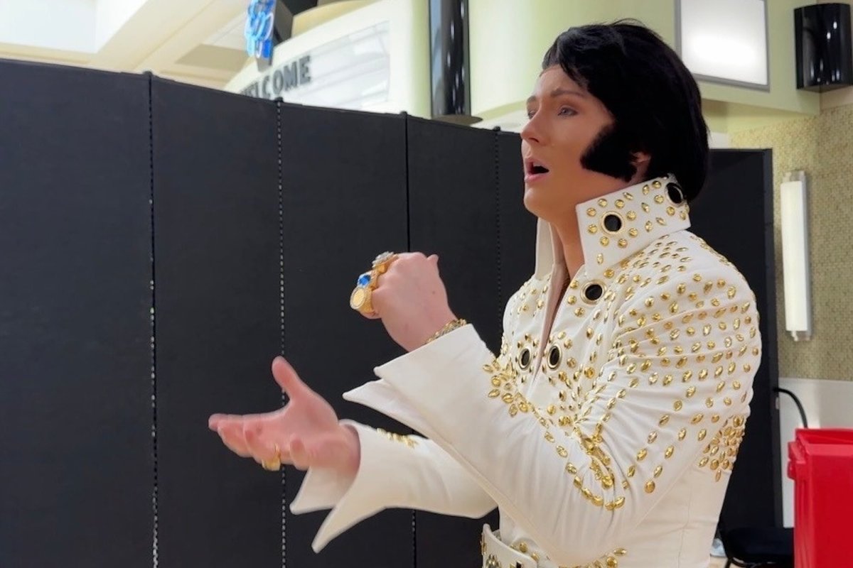 A young man dressed as Elvis Presley with a large black wig, sideburns and white jumpsuit practicing singing.