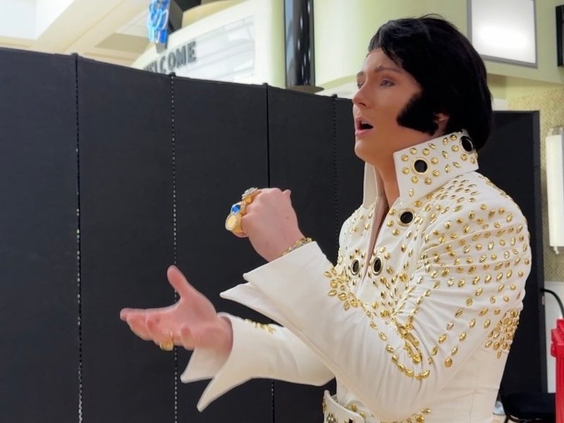 A young man dressed as Elvis Presley with a large black wig, sideburns and white jumpsuit practicing singing.