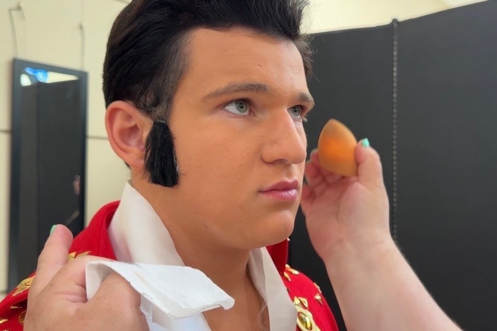 A young man dressed as Elvis Presley with a large black wig and sideburns has makeup applied to his face.